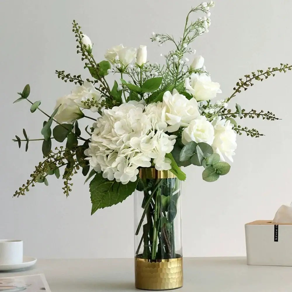 Vase en verre clair à base et bord martelés dorés avec hortensias, roses et verdure blancs